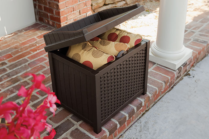 A brown deck box with patio furniture cushions inside sitting on a brick front porch
