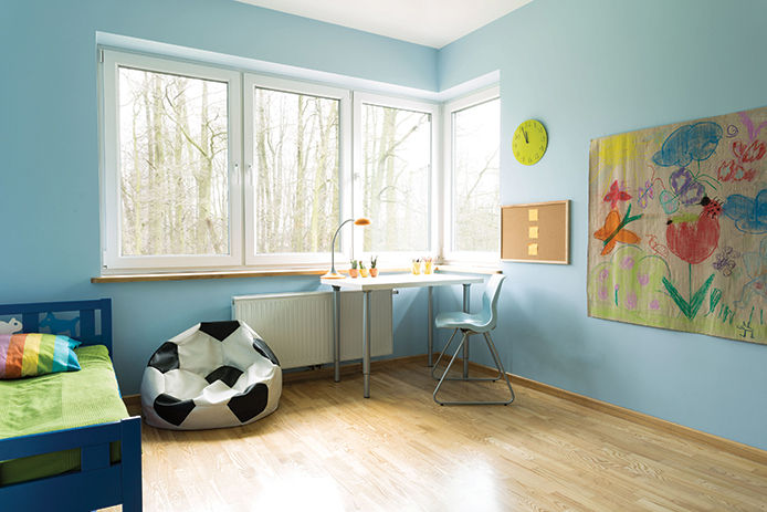 A childrens room with blue walls and four windows. There is a small wooden blue bed with a soccer bean bag and a small desk in the corner