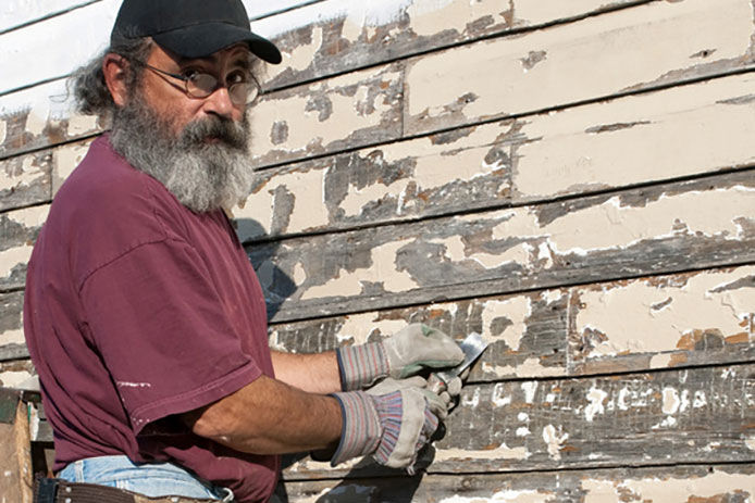 A older gentleman in a red shirt scraping paint off od wooden siding with a metal paint scraper on a sunny day. 