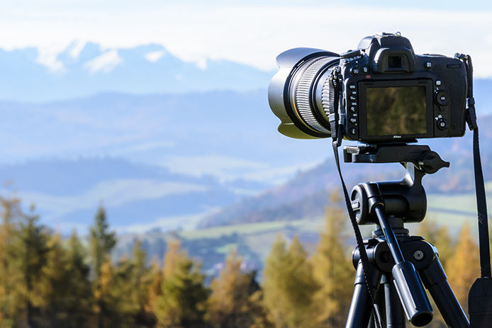 A professional black digital camera is mounted on a tripod overlooking a mountain and valley of green trees. 