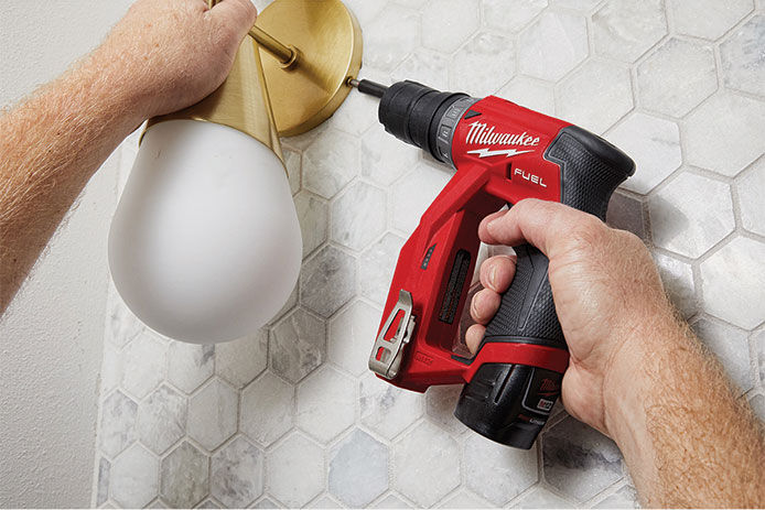 A hand holds a Milwaukee M12 FUEL Brushless 3/8 In. Installation Cordless Drill, fastening a brass wall sconce base to a tiled wall, with a glass light fixture and reflections visible below.