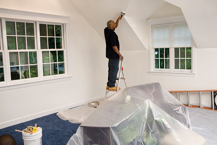 A man on a step ladder painting the ceiling with a canvas drop cloth covering the floor and furniture