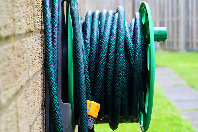 A green garden hose on a residentail garden wheel connected to a tan brick wall