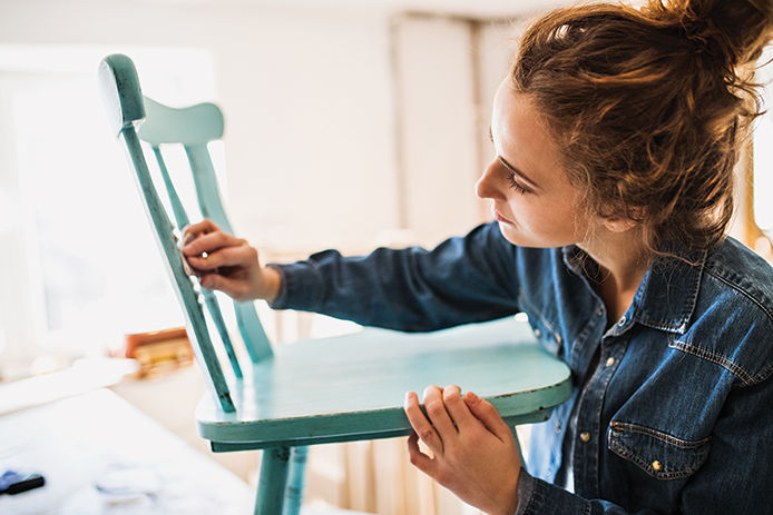 A young woman wearing a jean jacket sanding down a high gloss painted wooden chair 