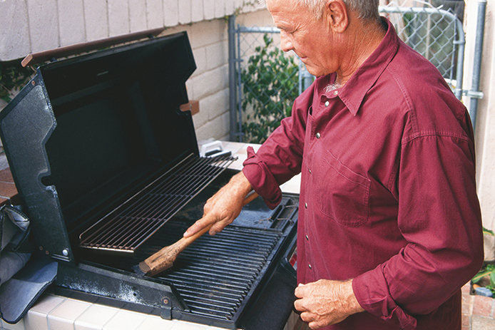Elderly man wearing a red shirt, standing by a gas grill in the snow-covered winter landscape, grilling food for a winter outdoor barbecue. The man is dressed warmly for the cold weather and is using cooking utensils to prepare food on the grill.