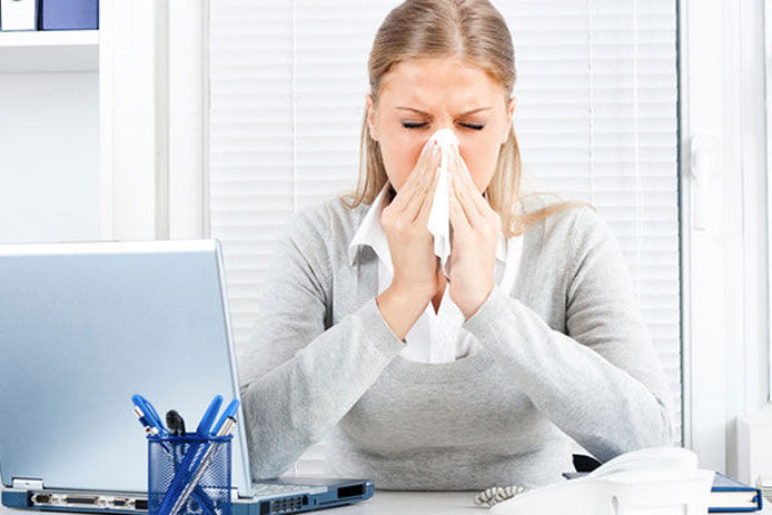 A woman sitting at her desk holding a tissue up to her nose sneezing