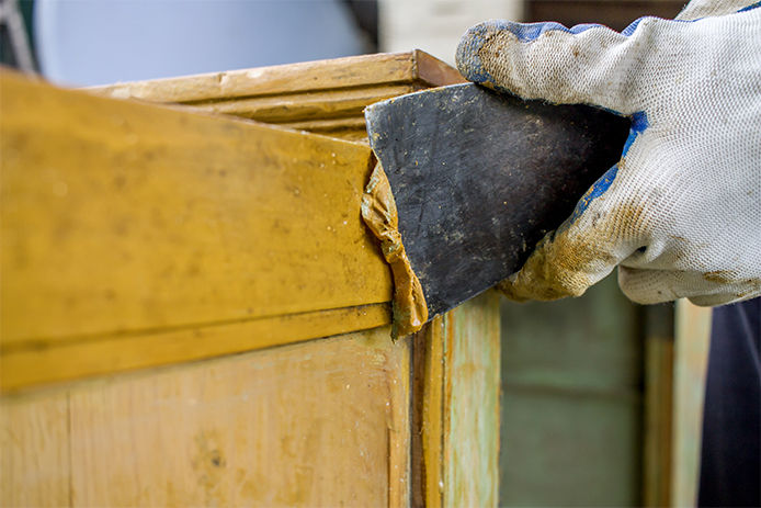 Close-up of a person using a paint scraper to remove yellow paint from wood