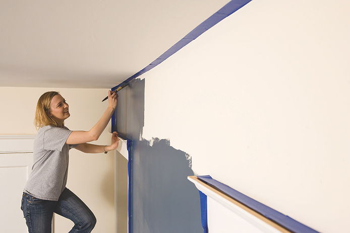 A young woman smiling while standing on a step ladder to cut out the top of a bedroom wall while painting 