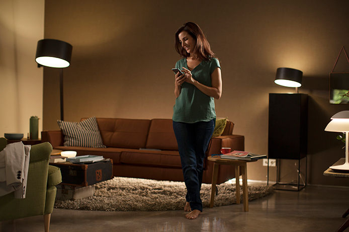 Woman standing in her living room looking at her phone with a couple of lamps turned on in a dimly lit room