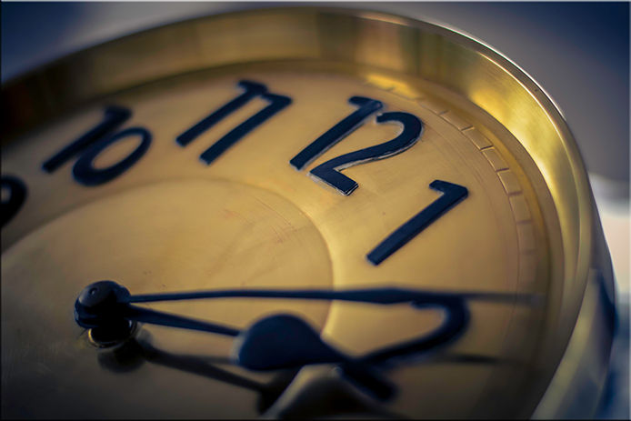 A closeup of a gold clock face against a grey background. The clock face features sans-serif numbers in black and black hands, showing the time 2:10. 