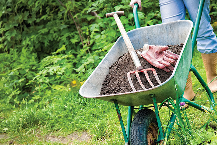 A close up of a woman pushing a green wheelbarrow filled with dirt, a pitch fork and garden gloves