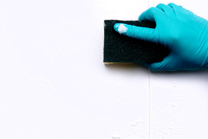 A close up image of a hand wearing a blue nylon glove holding a sponge wiping down a wall with wallpaper remover