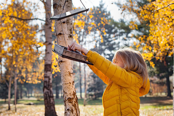 Young girl with blonde hair wearing a yellow jacket putting bird seed in a hanging bird feeder