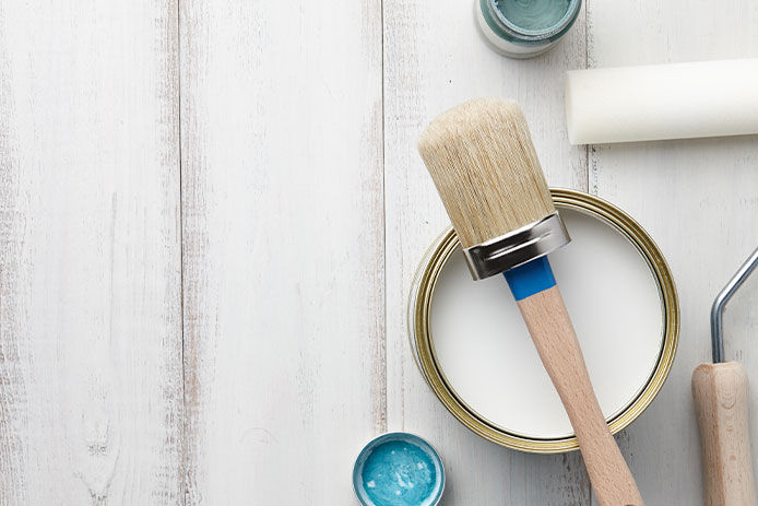 A tan bristled paint brush sits across the top of a can of white chalk paint. The can rests on a white-washed wooden surface next to a paint roller and smaller cans of turquoise-colored paint. 