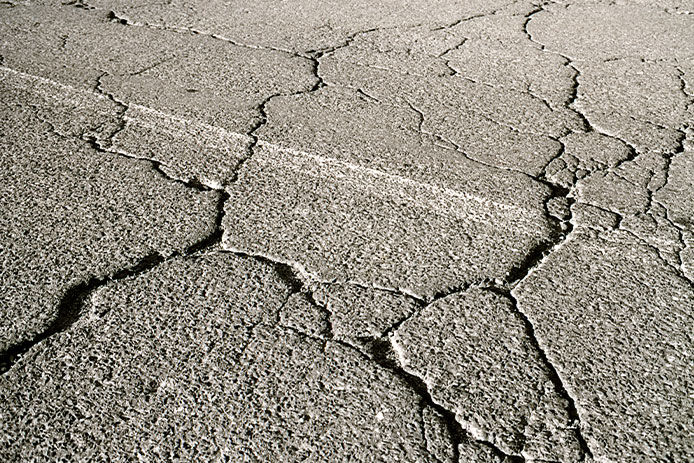 An asphalt surface shows signs of wear and tear, including significant fading to lighter grey and deep alligator skin-like cracks.