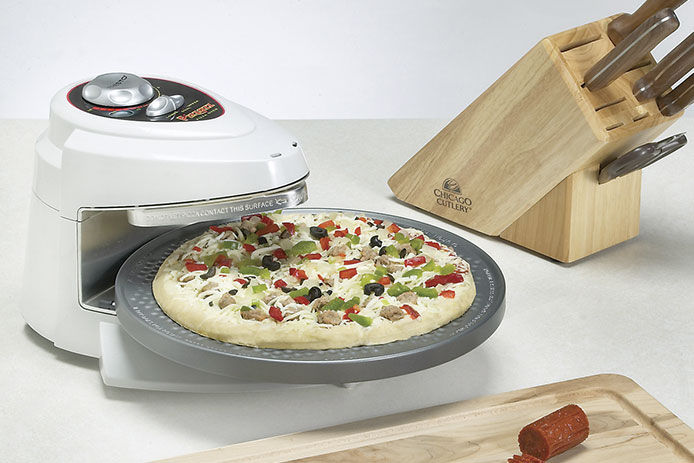 A white countertop pizza cooker is cooking a pizza on a white counter next to a knife block and a cutting board. The pizza has sausage, vegetables, and cheese toppings.