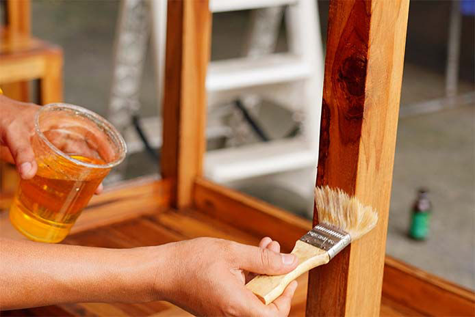 Person brushing stain on a piece of wood with a plastic cup of gel stain in the other hand