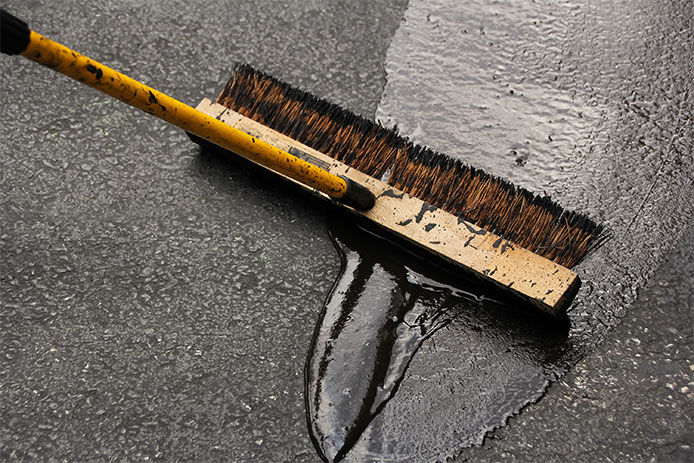 A long, flat driveway asphalt brush is shown atop fresh asphalt. The black asphalt has a wet glossy sheen, and the brown wooden brush shows flecks of the asphalt material.