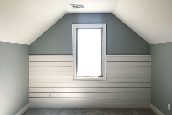 A barn shaped attic room that is accented with shiplap wainescotting. The walls are a seafoam blue color. The attic has a centered window that is shining light into the attic. 