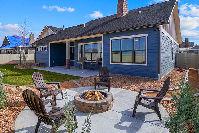 modern landscaped new construction home with blue sky and puffy white clouds. Covered back patio. Features new landscaping and a fire pit with chairs surrounding it.