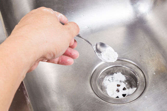 Person with baking soda on a spoon sprinkling into kitchen sink drain