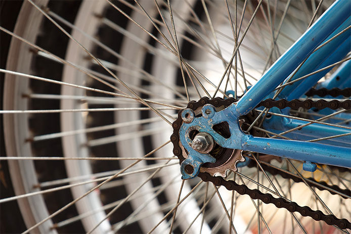 This is a close-up image of a bike tire spoke. The spoke is thin and made of silver metal, with a cylindrical shape and a slight curvature towards the center. It connects the black hub of the bike wheel to the outer rim, forming part of the wheel structure. 