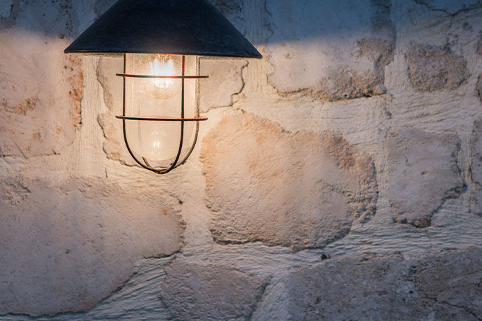 A glowing outdoor light fixture features a black wire cage surrounding a lightbulb with a flared fixture. There is a rock wall illuminated behind the fixture. 