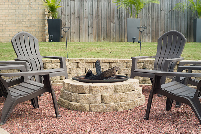 A paver brick firepit with four black adirondack chairs and tiki torches surrounding it 
