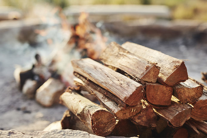 Medium-sized wooden logs are stacked in formation near a rock-surrounded fire pit, which is smoldering in the background.