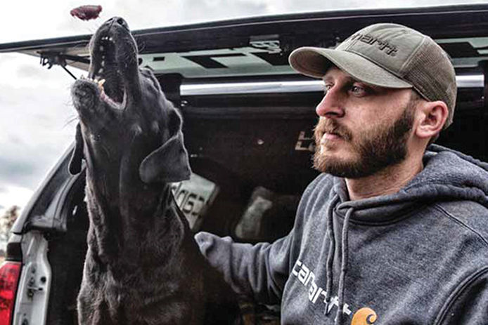 A man sitting on the tailgate of a truck with his black dog wearing Carhartt gear