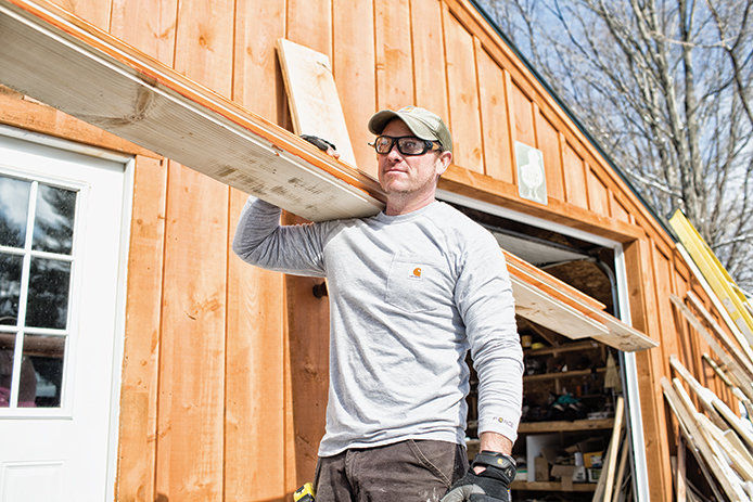 A middle aged man wearing a grey Carhartt long-sleeved shirt and carrying wooden boards