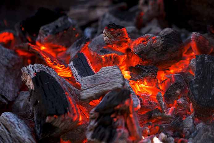 A closeup image of black and grey charcoal embers burning under a bright red-orange flame. 