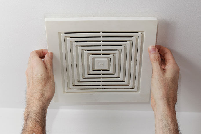 A person's hands hold onto either side of a square exhaust fan. The fan is white and mounted on a white ceiling.