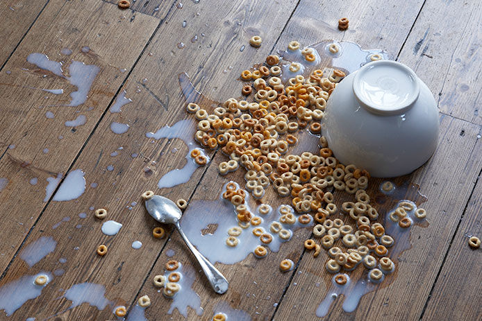 A bowl of spilled cereal all over wooden floor with bowl overturned