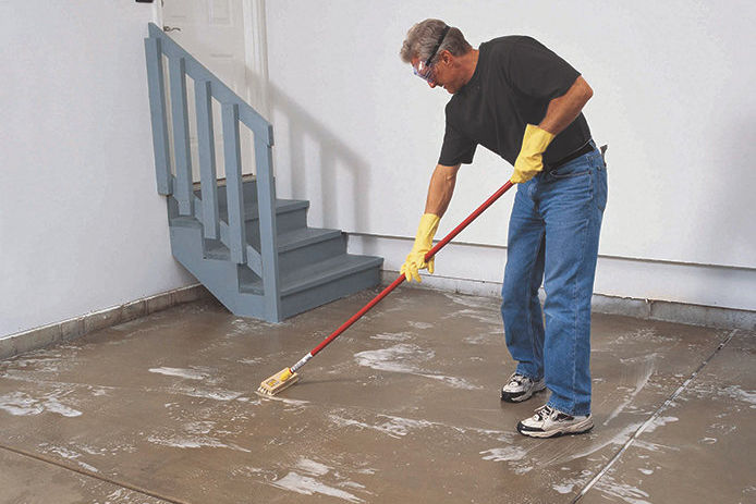 A middle aged man wearing jeans, a black t-shirt, and yellow rubber gloves preparing the surface of a garage floor by applying degreaser and etching