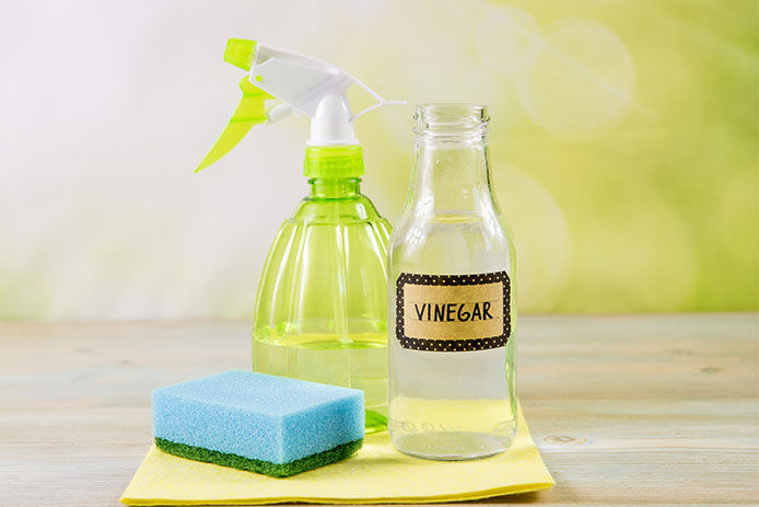 A lifestyle image of a spray bottle, blue and green sponge, and a glass bottle of vinegar to create a natural cleaning solution