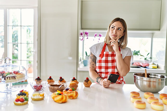 Woman with many baked goods on her kitchen countertop while she holds her smart phone in one hand and is thinking