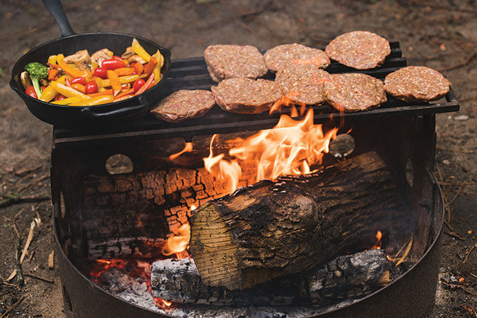 Skillet with veggies and a cooking grate with uncooked patties over the fire