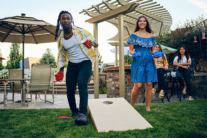 A group of friends playing a Cornhole bean bag toss game in a backyard of a home.