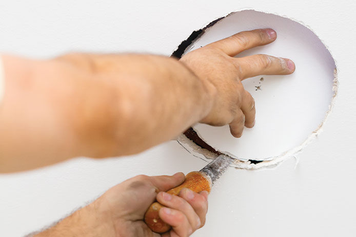 A person uses a small hand saw to cut a circular opening in white drywall.