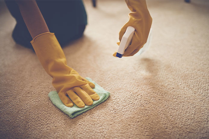 Woman spot cleaning her carpet with a spray bottle and microfiber cloth while wearing yellow rubber gloves