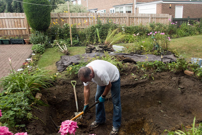 a man wearing a white shirt and jeans who is digging out a pond in his backyard. He is hunched over, using a shovel to dig out the pond as the soil is visible in front of him. The backyard is surrounded by pink flowers, green grass, bushes, and rocks, providing a beautiful and natural environment.
