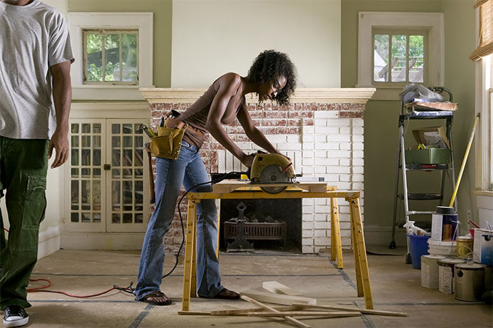 Woman using a power saw to cut a piece of wood in a living room that is being remodeled