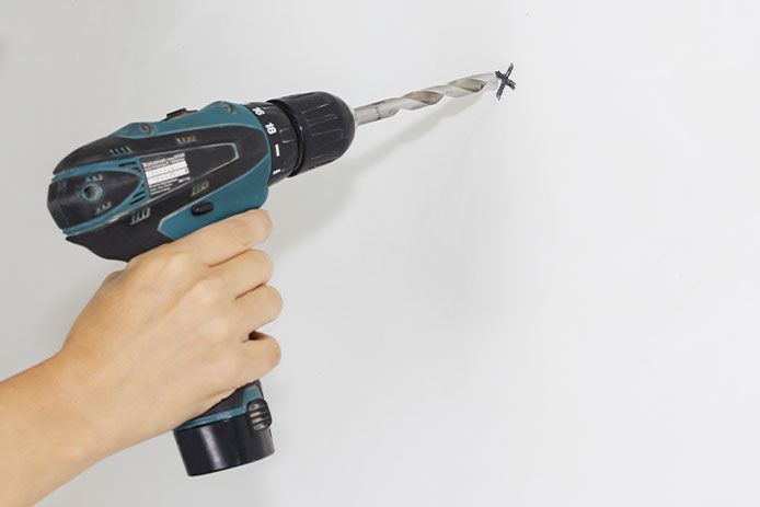 A person’s hand holds a blue cordless drill and touches the silver drill bit to a black X marking a spot on a white wall.