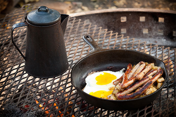 Eggs and sausages in skillet cooking on a grate over the fire with a kettle beside it