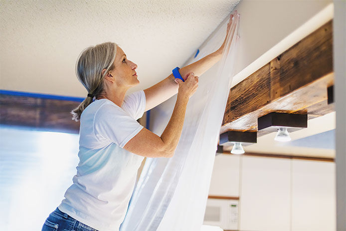 A woman standing on a step ladder hanging up plastic with painters tape to seal off a room 