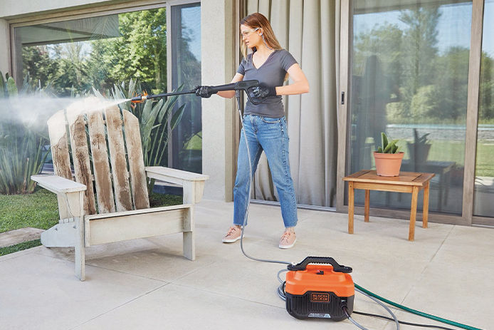 A young woman washing off her wooden adirondak chair with a electric pressure washer on her back patio