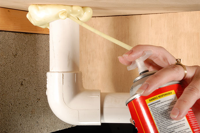 A woman holding a can of spray foam with the nozzle pointed at plumbing fixtures on a bathroom sink. 