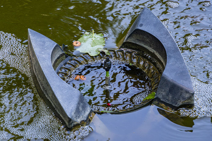a pond filter aeration system sitting on top of the pond water with a middle fountain spout. The focus is on the aeration system and the fountain spout, which is visible in the center of the image. 
