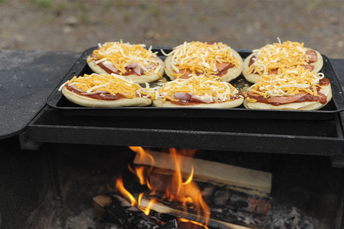 Six personal-sized cheese pizzas are shown cooking on a grill pan over an open fire.